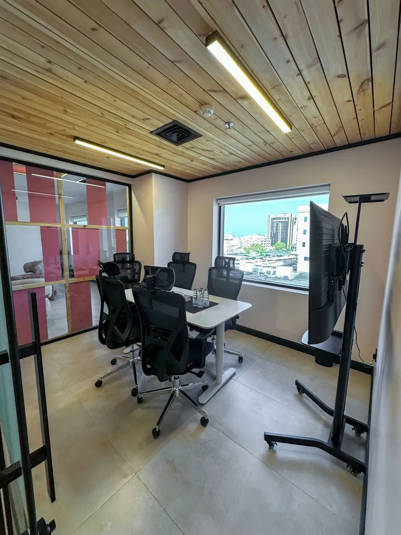 Black mesh chairs around a white table, a screen on a mobile stand and a city-view window